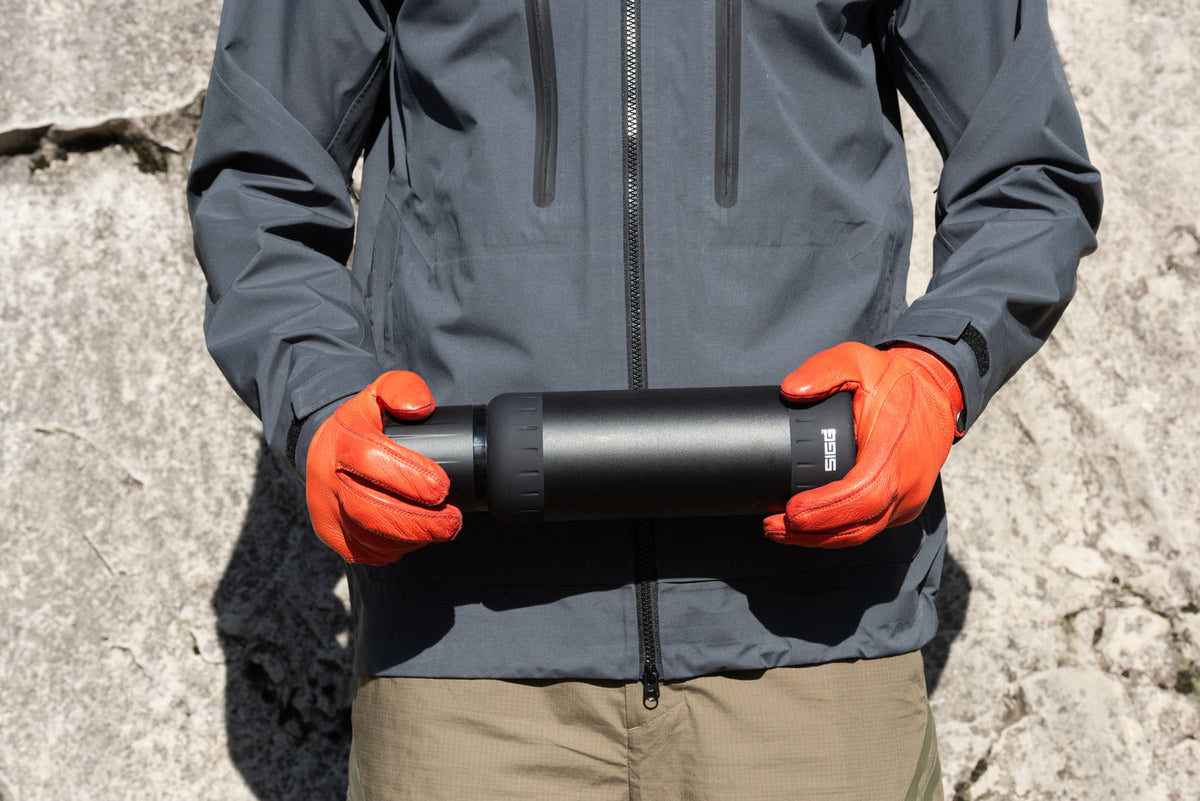 En person i grå skaljacka och orange handskar håller en svart termos utomhus